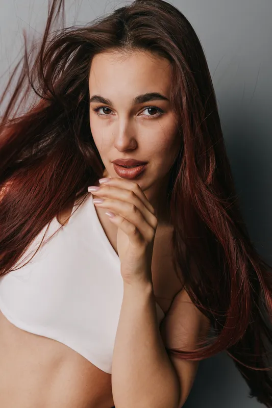 Young woman with long brown hair and natural makeup posing in white top against gray background. Closeup portrait with confident expression and hand near face in studio lighting.