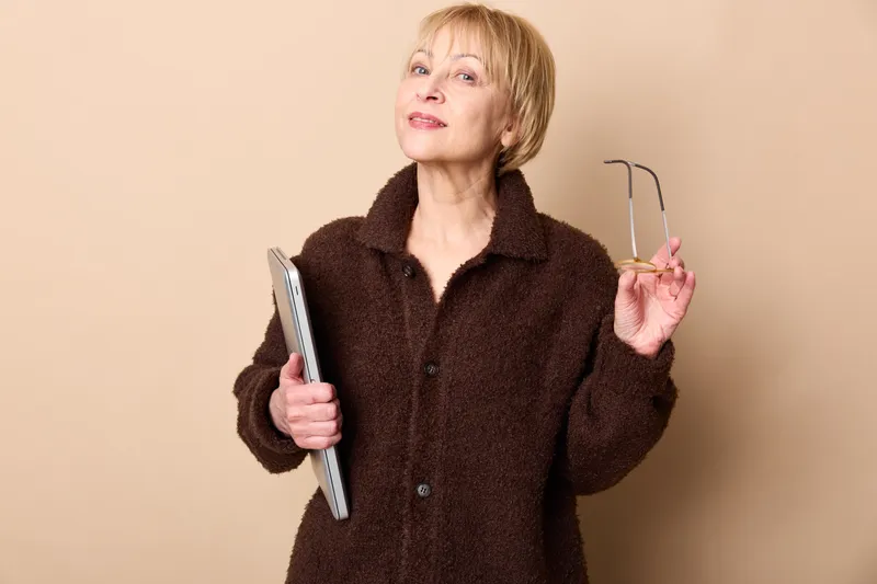 Woman senior holding notebook and eyeglasses in studio, beige background, thoughtful face, casual coat, blonde hair, middle age female, focused pose, calm mood, copy space
