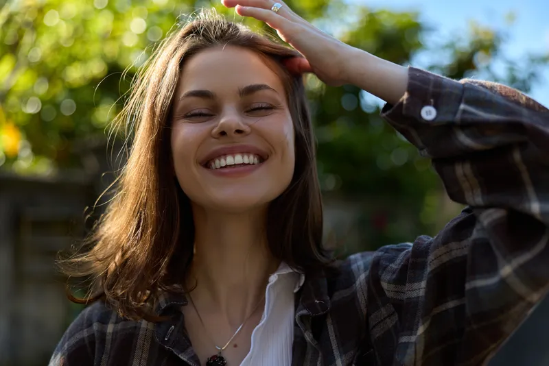 A cheerful woman enjoys a sunny outdoor moment, smiling with her eyes closed, hair softly blown by a gentle breeze. A candid lifestyle portrait celebrating joy and freedom
