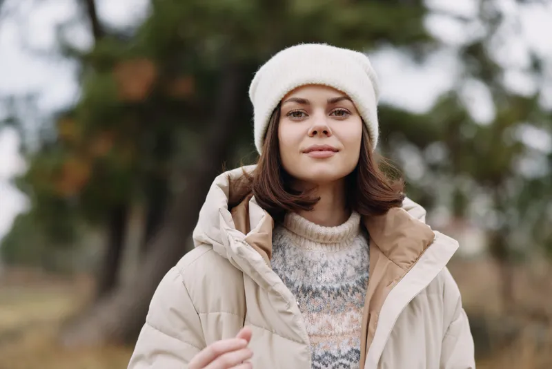 Winter portrait of a woman in a beige coat and knit sweater, wearing a white beanie, standing outdoors with blurred trees, conveying cozy seasonal fashion and calm mood