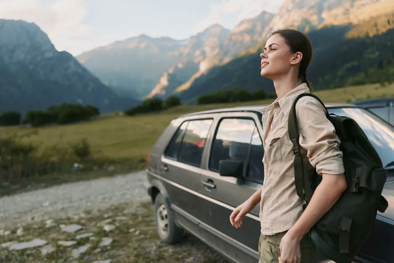 Young woman with a backpack walks beside a parked car in a rugged mountain valley, enjoying a bright crisp outdoor moment.