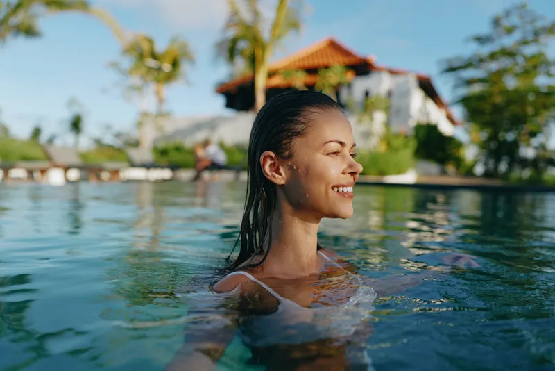 Cheerful woman in a sunlit pool, smiling with eyes closed, tropical resort backdrop, refreshing water, casual swimwear, relaxed mood, vibrant colors
