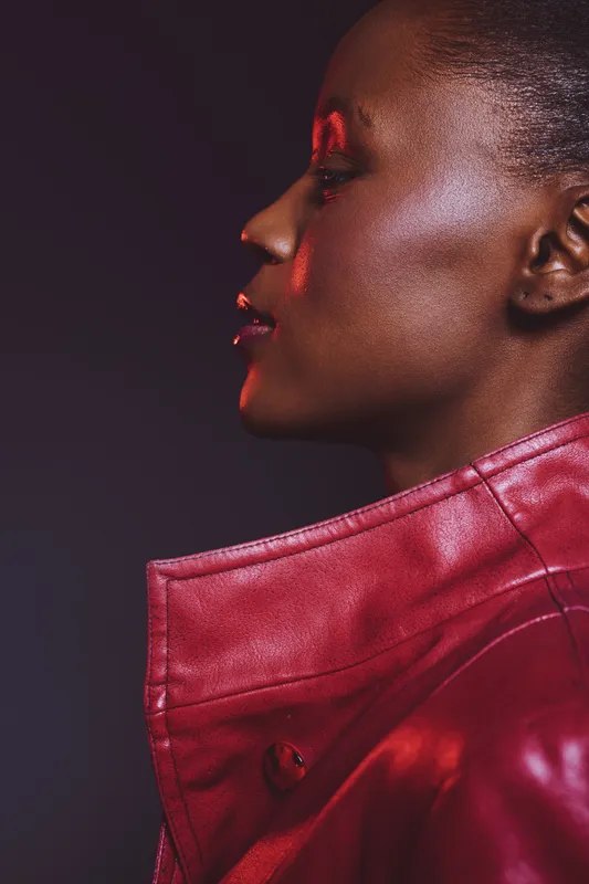 Profile portrait of confident dark skinned African woman wearing red leather jacket with high collar. Studio lighting highlights smooth skin texture and fashionable urban style, isolated on dark