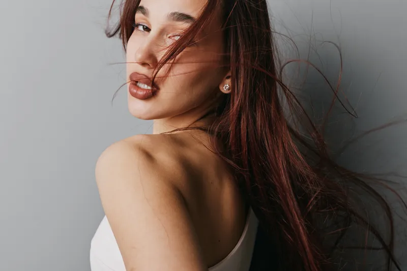 Young woman with long hair looking over shoulder with wind blowing strands, wearing white top against neutral background. Beauty, portrait and fashion concept with natural makeup and earrings.