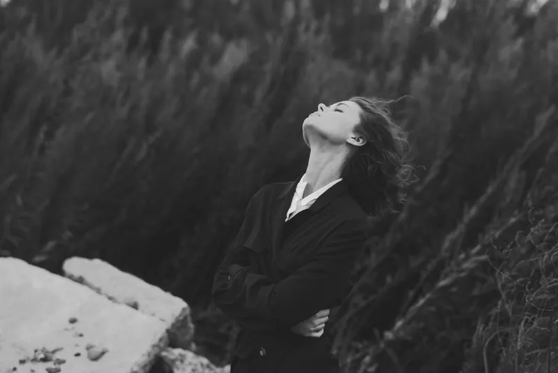 Contemplative woman gazing upward in a monochrome outdoor landscape, arms crossed, wind blowing through hair, solitary moment by rugged rocks, a moody study of resilience and nature