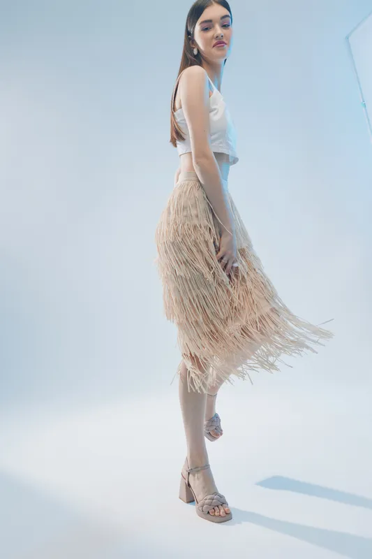 Young woman in a stylish fringed skirt and white top poses elegantly in a bright studio. Concept of woman beauty fashion with modern outfit and confident attitude.