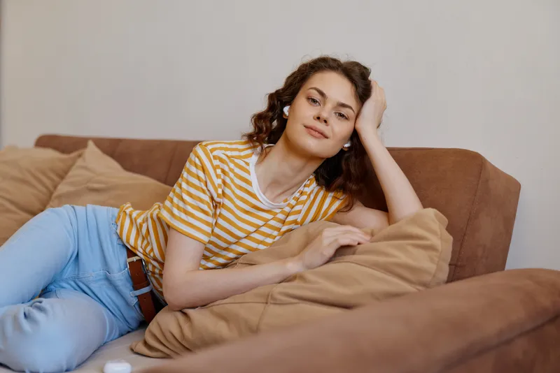 Young woman with curly hair lounges on a cozy sofa at home, resting her head on a soft pillow and gazing with a calm, content expression, softly dressed in casual stripes, creating a tranquil,