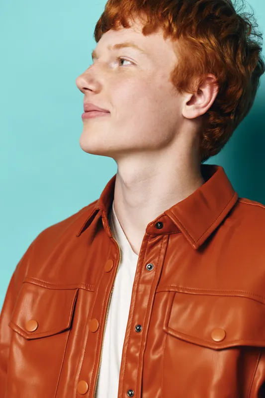 Young man with red hair wearing brown leather jacket and white shirt looking sideways with confident expression on blue studio background in closeup portrait.