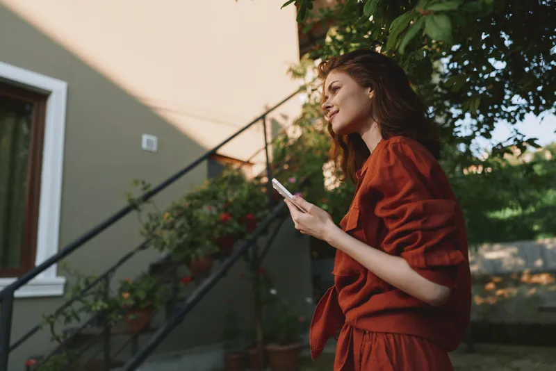 A woman in red stands on outdoor stairs, looking at her phone with a relaxed expression. A casual lifestyle moment in bright daylight