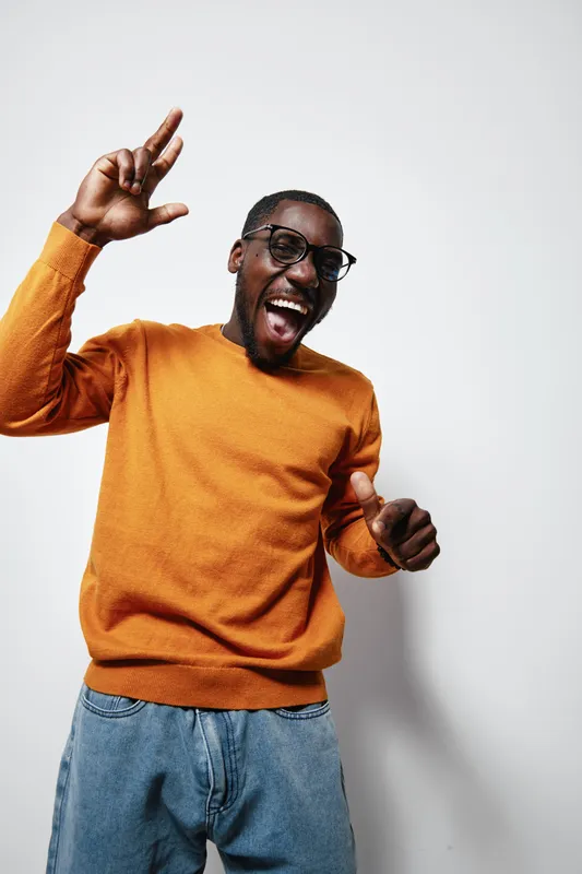 African American man with dark skin wearing glasses and orange sweater smiling and dancing with raised hands against white background in casual style.