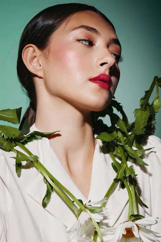 Close-up of a model with glossy makeup and red lips posing with green leaves. Creative beauty fashion concept with sharp facial features and soft studio lighting.