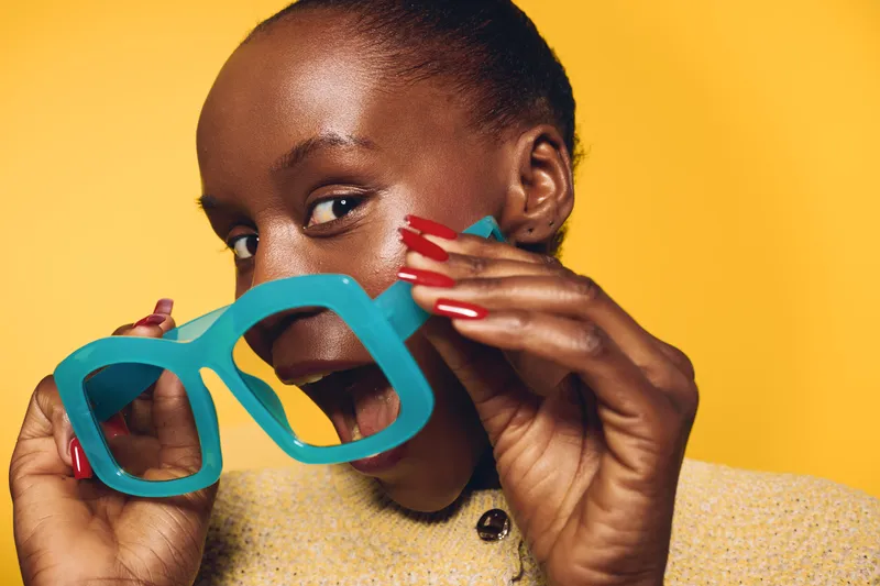 Happy African woman with dark skin holding large blue glasses in front of her face, smiling playfully on yellow background. Concept of joy, fun, and vibrant lifestyle.