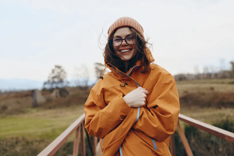 Woman lifestyle outdoors, smiling in yellow coat and knit beanie, wearing glasses, breezy rural field path, golden sunlight and soft wind, relaxed pose adjusting jacket, natural colors