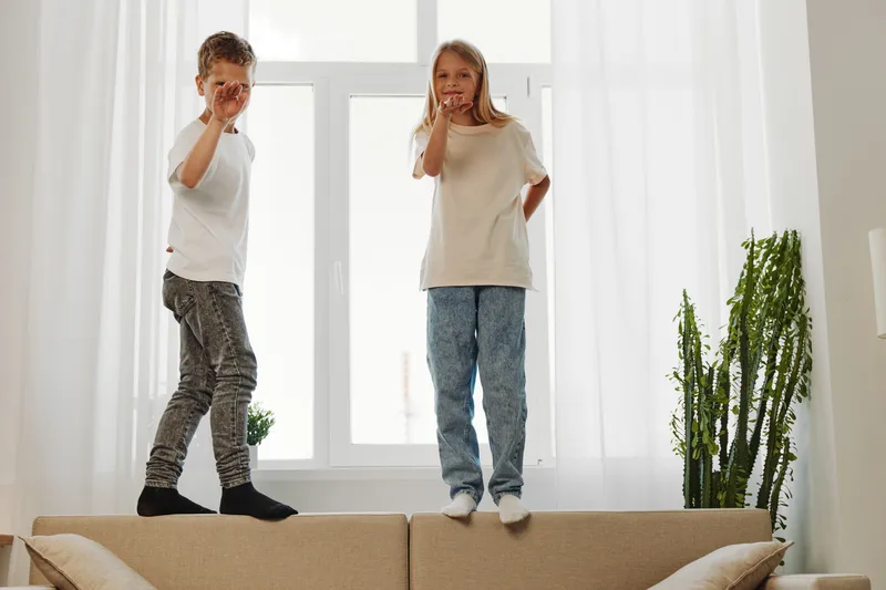Two happy children jumping on beige sofa in bright room with large windows, enjoying playful moment and active indoor fun at home with natural light.