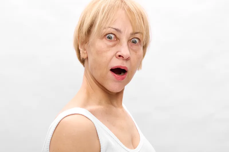 Surprised woman with short blond hair, wide mouth, shock expression, close face view, studio portrait, white background, sudden surprise emotion, soft studio lighting