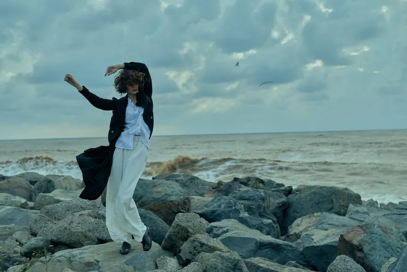 Woman coat wind ocean rocks movement on stormy cloudy seaside with dynamic posing windblown gesture fashion mood outdoor photography capturing dramatic breeze amid gray horizon and rugged coast nature