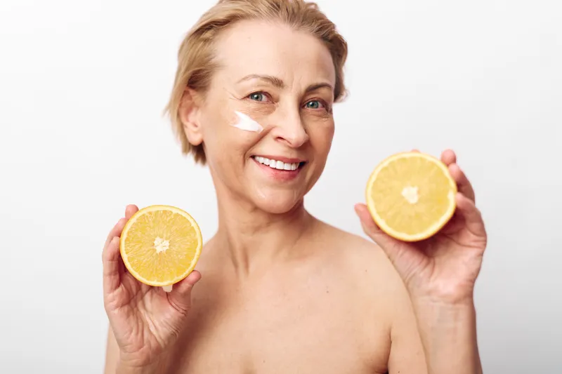 Smiling mature woman holds two halves of fresh orange near face with cream on cheek. Concept of natural skincare, healthy lifestyle, anti aging, vitamin c, and radiant skin care routine.