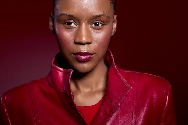 Close-up portrait of a confident black woman with dark skin wearing a red leather jacket and red top. Studio shot with dark red background. Concept of fashion, beauty, and empowerment.