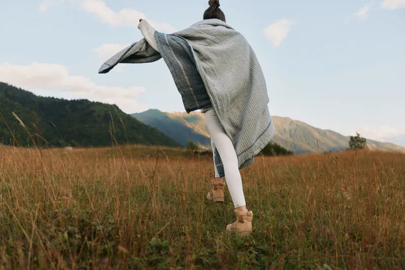 Woman lifestyle outdoors walking in grassy field wrapped in cozy blanket with boots on, scenic mountain background under clear sky, autumn atmosphere