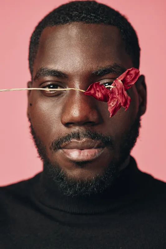 Creative portrait of African man with dark skin wearing black turtleneck holding red flower on eye against pink background in conceptual visual style for artistic expression.