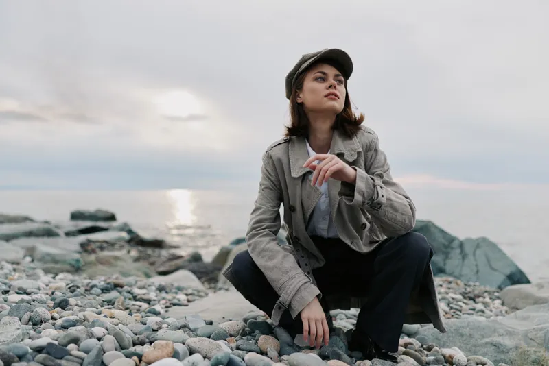 Young woman in casual clothes and cap squats on rocky beach during cloudy sunset. She looks away thoughtfully, capturing mood of relaxation, contemplation, and nature outdoors.