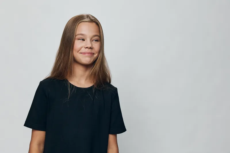 Girl with long brown hair and black t shirt smiling and looking to side on plain light background in studio portrait. Youth, casual clothing and happiness with natural expression.