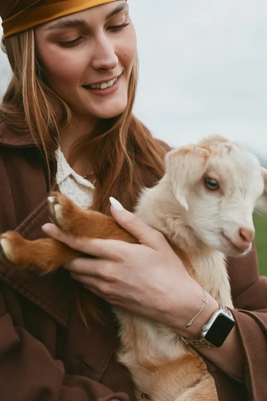 Young woman gently holds a baby goat in her arms while smiling softly outdoors in a cozy autumn outfit with a beanie and smartwatch on her wrist