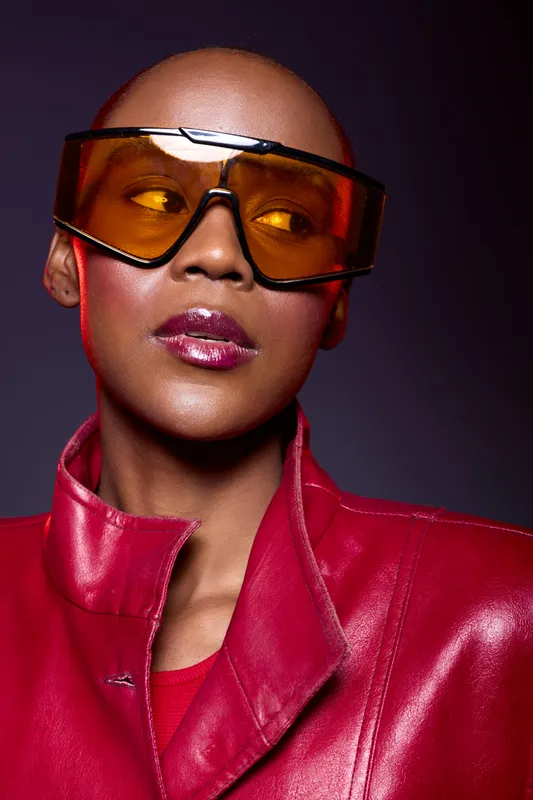 Portrait of a confident black woman with dark skin wearing large orange sunglasses and a red leather jacket. Studio lighting highlights her glossy lips and smooth bald head with a purple background.