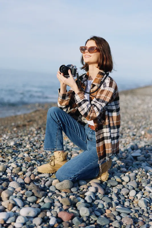 Female photographer on a rocky beach, kneeling with a camera, wearing a plaid shirt, jeans and boots, capturing seaside scenery in bright daylight and wind.