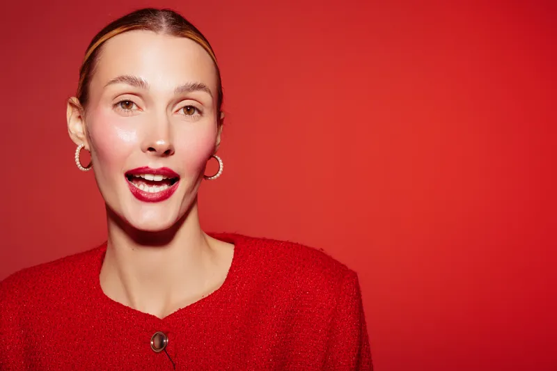 Portrait of a smiling woman with slicked back hair wearing red clothes and hoop earrings on red background. Concept of fashion, beauty, confidence, and vibrant style.