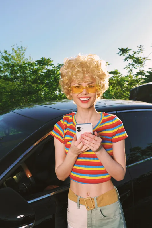 Young woman lifestyle shot with retro film color, smiling while using a phone beside a sleek car, wearing a bright striped crop top and sunglasses in a sunny outdoor scene