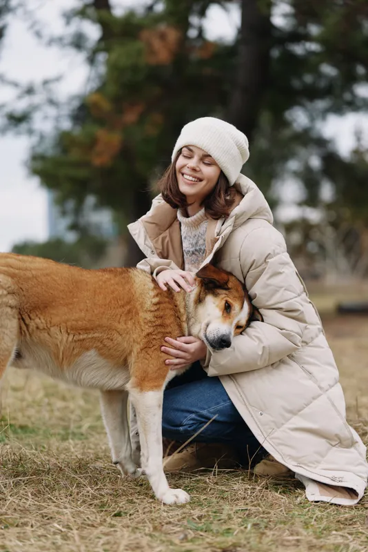 Woman in a warm winter coat hugs a dog outdoors, smiling with joy as autumn leaves fall. A tender scene of companionship and affection in a quiet park setting, natural and calm atmosphere.