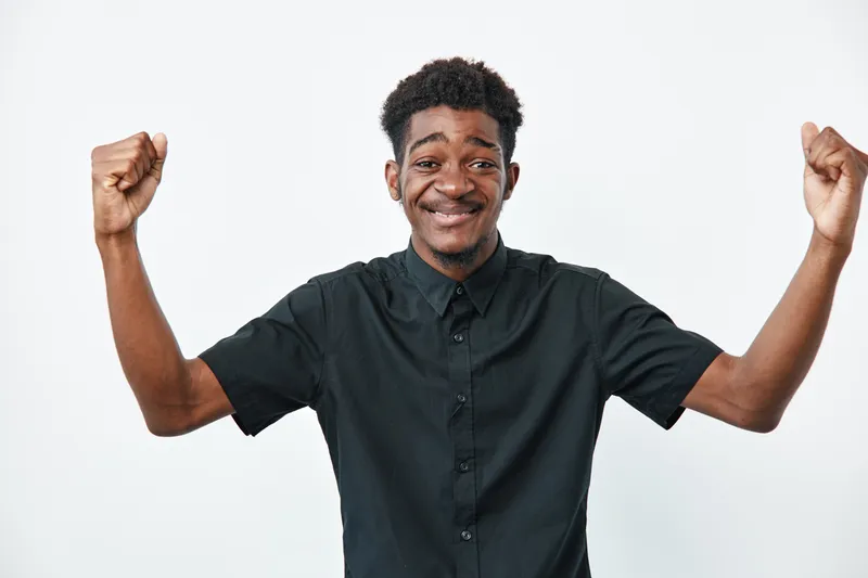 Young black man with dark skin wearing black shirt smiling and raising fists in celebration on white background. Happy African male expressing success and joy with confident pose.