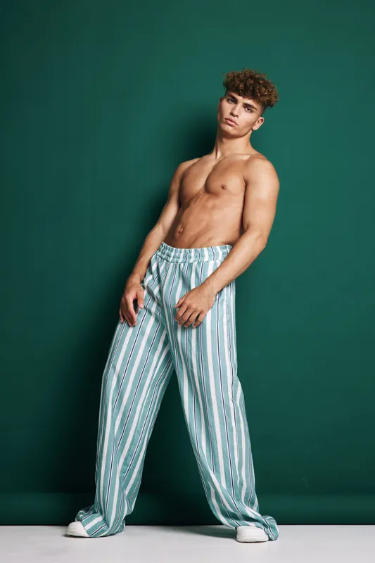 Young man with muscular body standing against green background wearing striped loose pants and white sneakers with confident pose looking at camera at studio.
