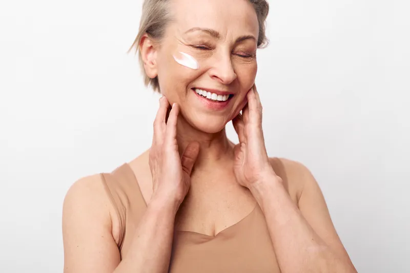 Smiling mature woman with closed eyes applies face cream on cheek, touching neck gently. Concept of skincare, natural beauty, self-care, anti-aging and wellness in studio with soft lighting.