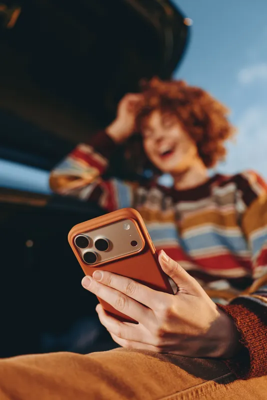 Woman lifestyle in car interior, hand holding smartphone with brown case, child sitting nearby smiling during travel, daylight, blurred background, casual leisure moment, closeup view