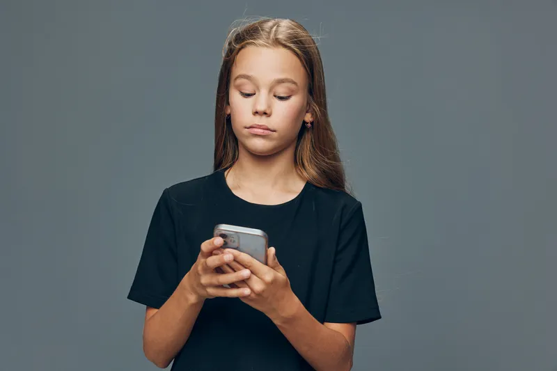 Girl wearing black shirt holding smartphone and looking down with serious face on grey background. Child with long hair using mobile device for text or app in studio.
