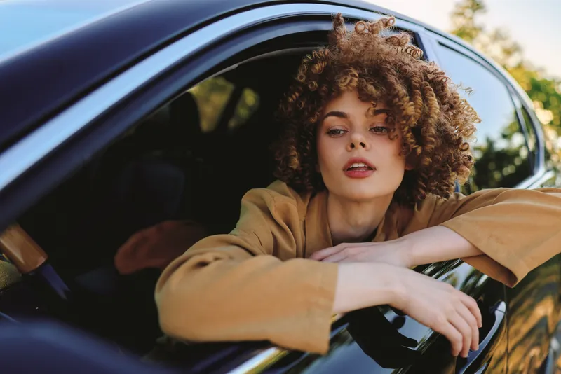 Young woman with curly hair leans from a car window in a casual brown jacket, enjoying a sunny drive and a carefree street style moment, capturing a spontaneous mood.