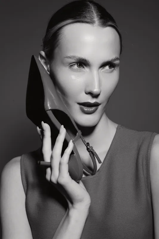 White woman with light skin holds a high heel shoe near her face, looking to the side. Black and white studio portrait with elegant stylish concept.
