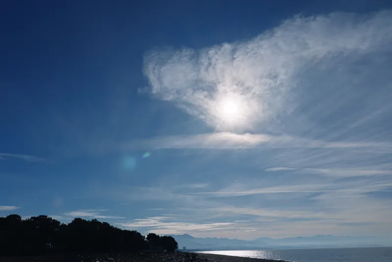 Serene sea and coastline under a bright sun, calm blue sky with wispy clouds, a distant horizon reflecting on the water for a peaceful coastal landscape