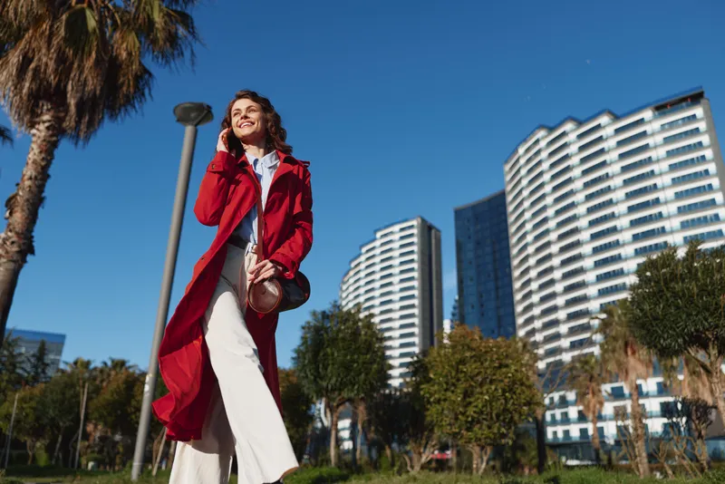 Young woman wearing red coat walking outdoors in city park with modern buildings and palm trees smiling and enjoying sunny day with positive mood and casual style.