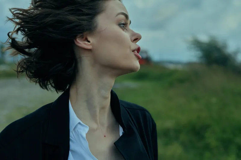 Profile of a woman in a blazer and white shirt standing in a windy outdoor field, conveying professional style, determination, and dynamic movement in a natural setting.