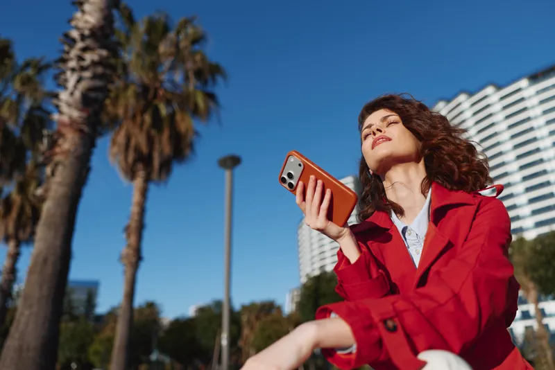 Smiling woman in a red jacket holding a smartphone outdoors in the city, looking up with a confident pose. Sunny day, urban palm trees and buildings, portrait.