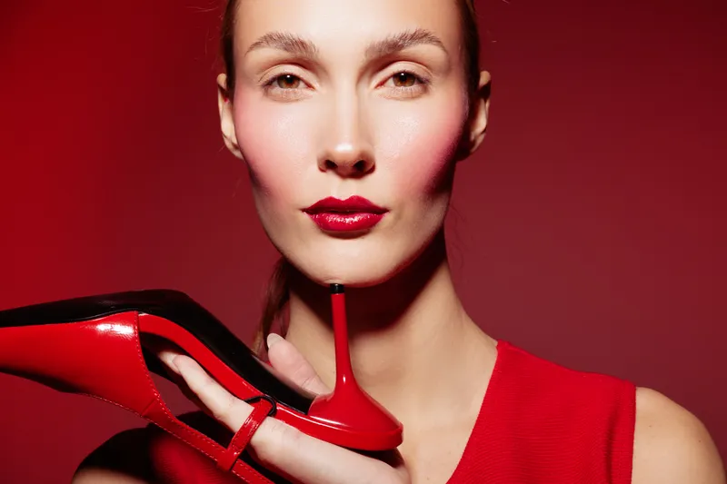 Close-up of a woman with red lipstick holding a red high heel shoe near her face. Red background and red dress create a bold fashion concept with strong color contrast and style.