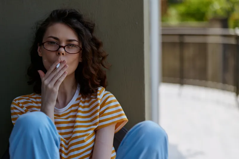 A thoughtful woman in a striped yellow and white shirt sits indoors, knees drawn up, exhaling softly. A calm home setting conveys casual, reflective mood and modern lifestyle for wellness and self