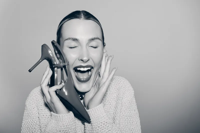 Happy Caucasian woman with light skin holds red high heel shoe near face, eyes closed and mouth open. Concept of fashion, style, joy, and female expression in studio.