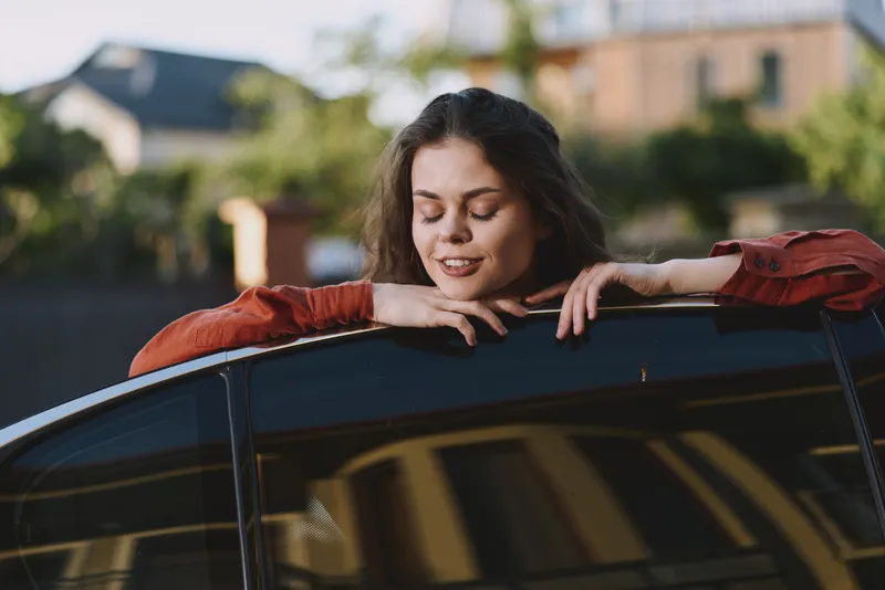 Young woman rests on a car window ledge, smiling softly in a sunlit outdoor setting with a relaxed pose and a warm breeze guiding the red blouse