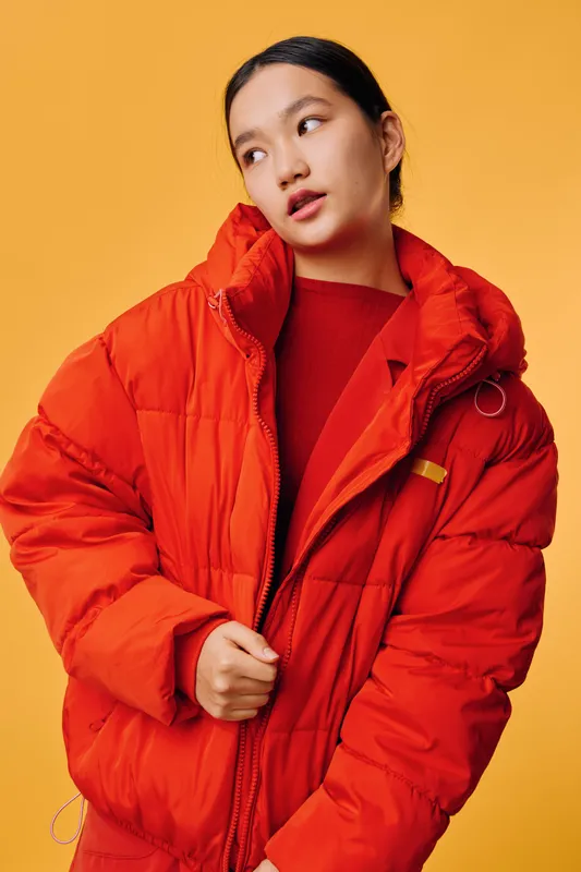 Woman wearing bright red puffer jacket and sweater posing against yellow background. Stylish Asian female model with natural makeup in casual warm winter clothing at studio.