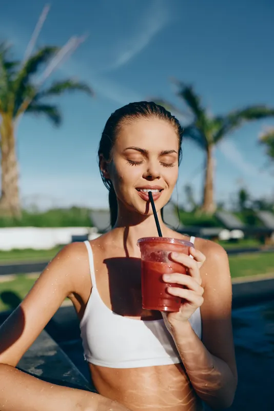 A relaxed young woman in a white bikini top sips a red smoothie through a straw by a bright pool outdoors, palm trees in the background. A lifestyle moment of leisure and sunny warmth.