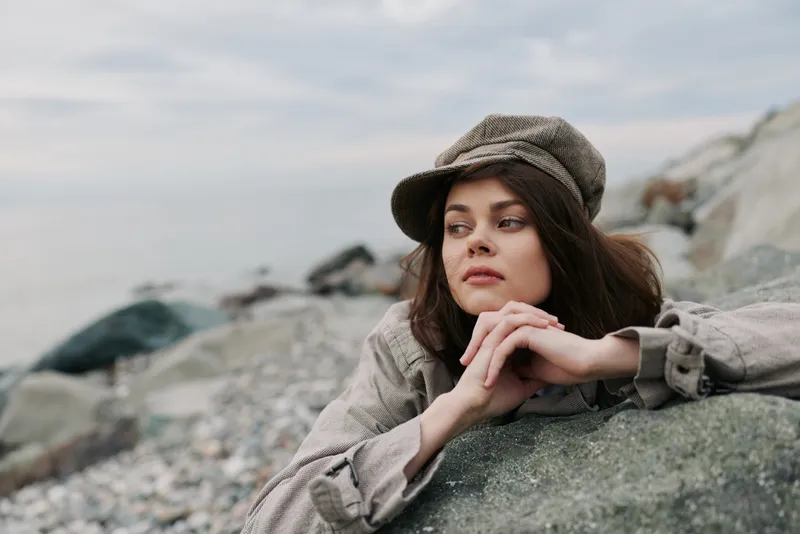 Young woman in a knitted hat and jacket leans on a rock at the rocky beach. She looks away thoughtfully under a cloudy sky. Concept of contemplation and nature outdoors.